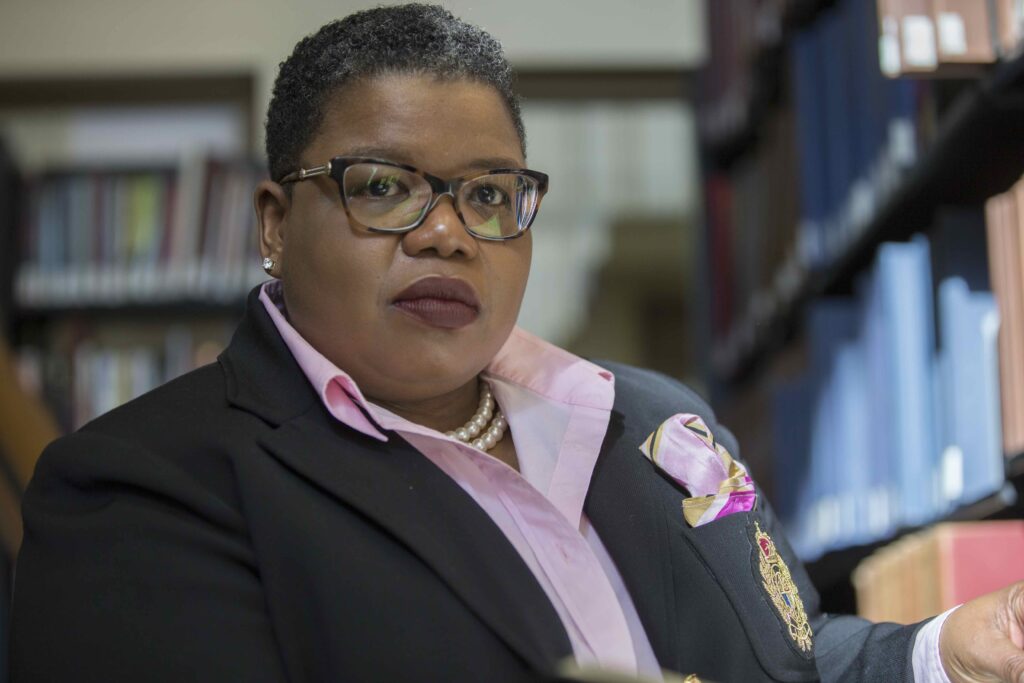 A person wearing glasses, a black blazer with a pink pocket square, and a pearl necklace sits in a library, looking directly at the camera with a serious expression. Bookshelves are visible in the background.