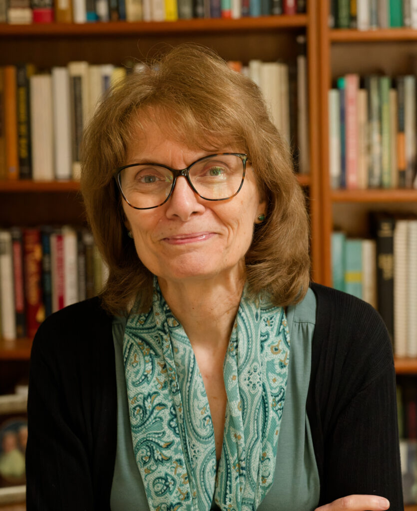 A woman with glasses and shoulder-length hair smiles softly, wearing a green top and patterned scarf, standing in front of bookshelves filled with books.