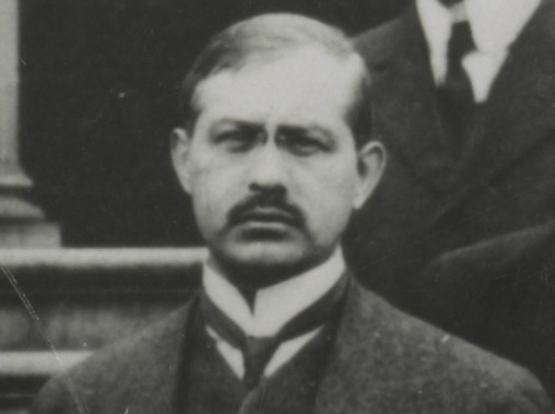 A man with short hair and a mustache, wearing a suit and tie, looks directly at the camera with a serious expression. The photo is black and white and appears historical.