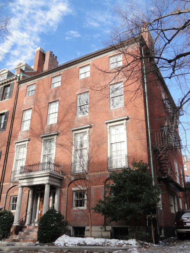 A tall, red-brick townhouse with white trim, large windows, a columned entrance, an iron balcony, and an external fire escape. Bare trees and patches of snow are visible in front.