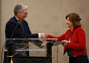A man in a suit and a woman in a red jacket smile and shake hands across a podium during an indoor event, with the woman holding a certificate.