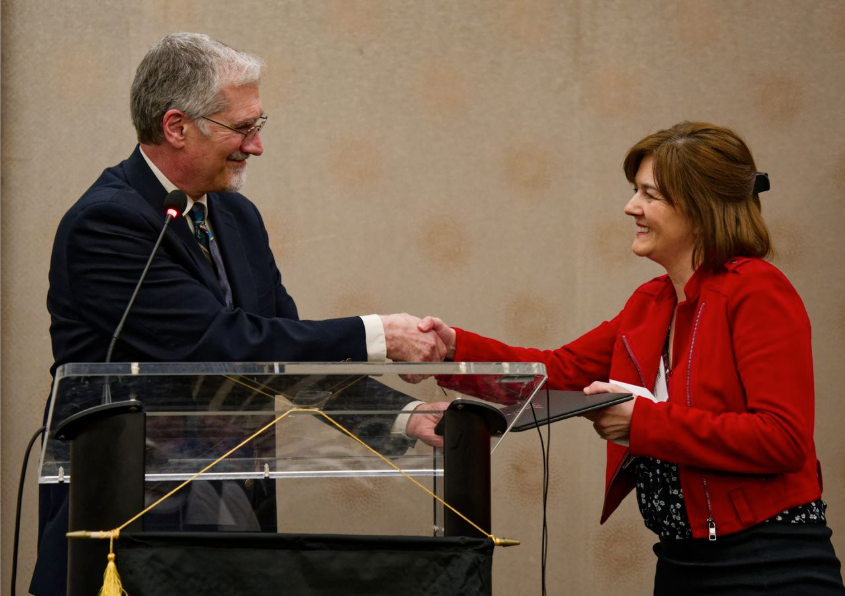 A man in a suit and a woman in a red jacket smile and shake hands across a podium during an indoor event, with the woman holding a certificate.