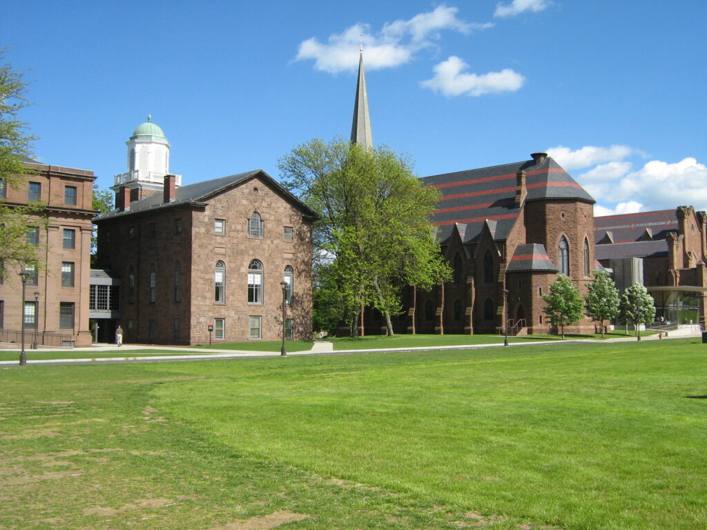 A college campus with historic brick buildings, a chapel with a tall steeple, and a large grassy lawn under a blue sky with scattered clouds.