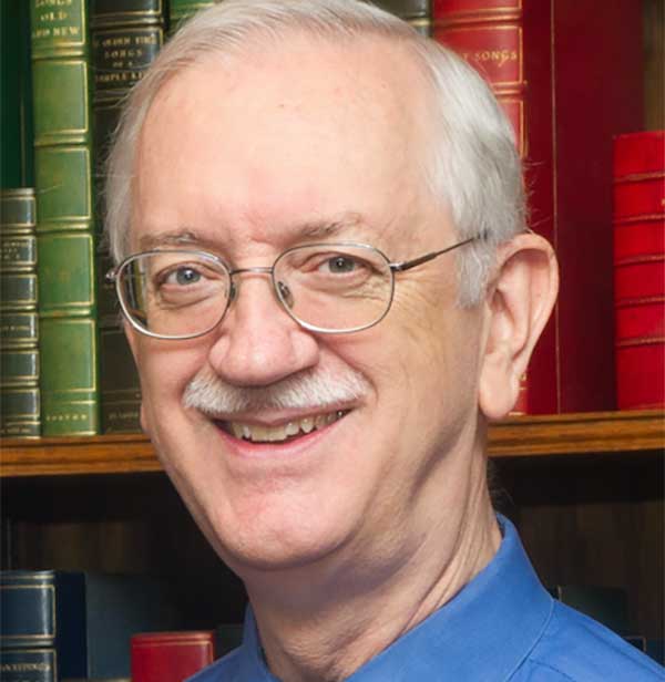 Smiling older man with glasses and white hair, wearing a blue shirt, standing in front of bookshelves filled with colorful books.