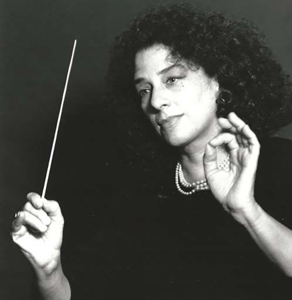 A woman with curly hair conducting with a baton, wearing a dark top and a pearl necklace, raises one hand while making a gesture with the other. Black and white photo with a dark background.