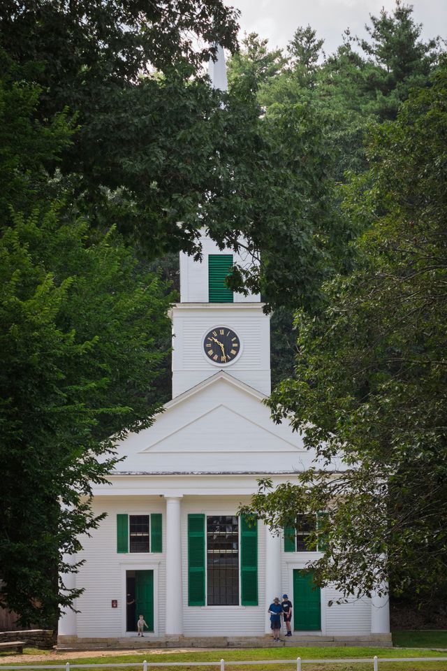 A white church with green shutters and a clock tower is surrounded by trees. Two people stand outside the front entrance.