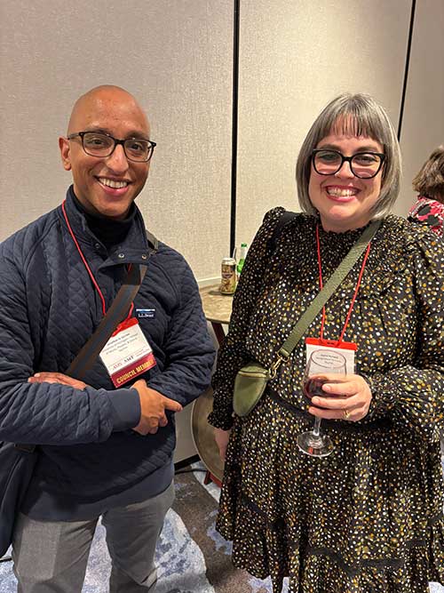 Two people smiling at an indoor event; one holds a drink and wears glasses and a patterned dress, the other wears a dark jacket and a conference badge.
