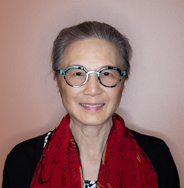 An older woman with gray hair in a bun, wearing round glasses and a red scarf with black script, smiles at the camera against a peach-colored wall.