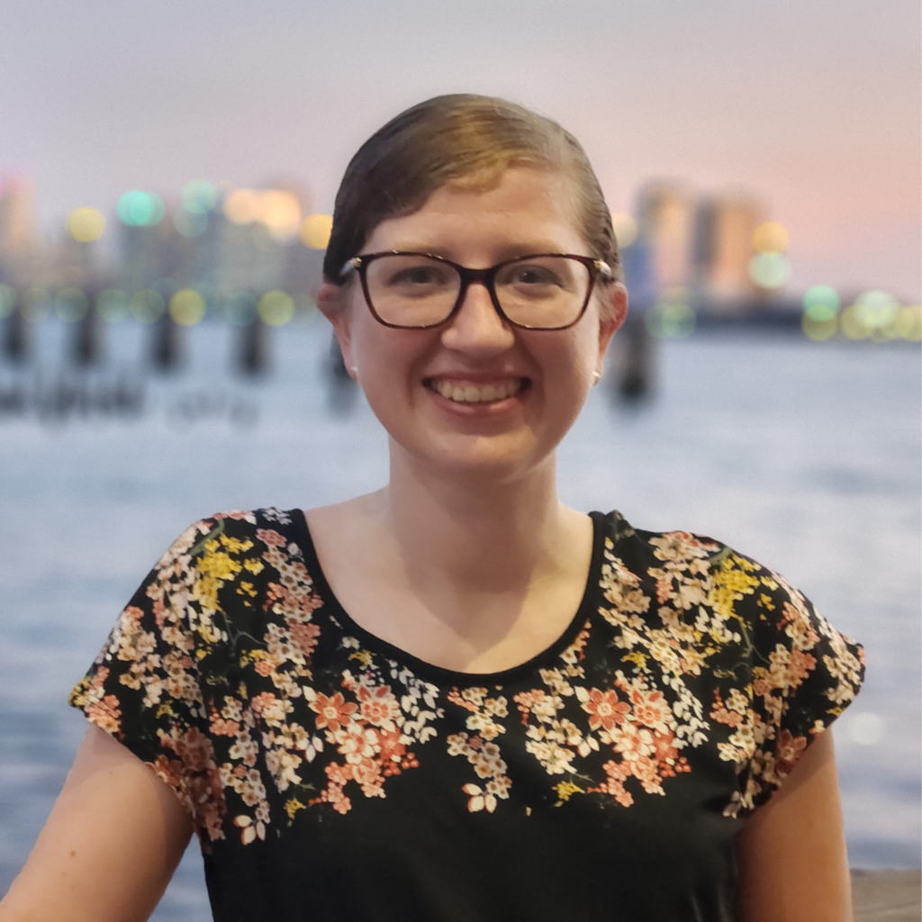 A smiling person with glasses and brown hair, wearing a black floral top, stands in front of a blurred cityscape and water at sunset.