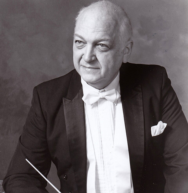 An older man in a tuxedo with a white bow tie and pocket square sits holding a conductors baton, looking slightly to the side and smiling softly.