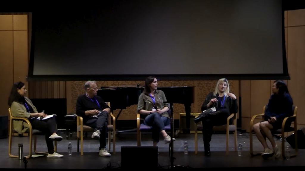 Five people sit in chairs on stage, engaged in a panel discussion. A piano and a large screen are visible in the background. One person gestures while speaking, and the others are listening. Bottled water is on the floor in front of them.