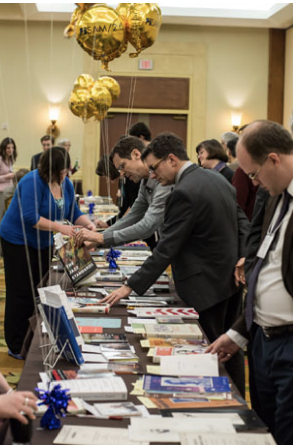 People at a conference browse a long table filled with brochures, pamphlets, and books. Gold balloons float above the table, and attendees are engaged in reading and picking up materials.
