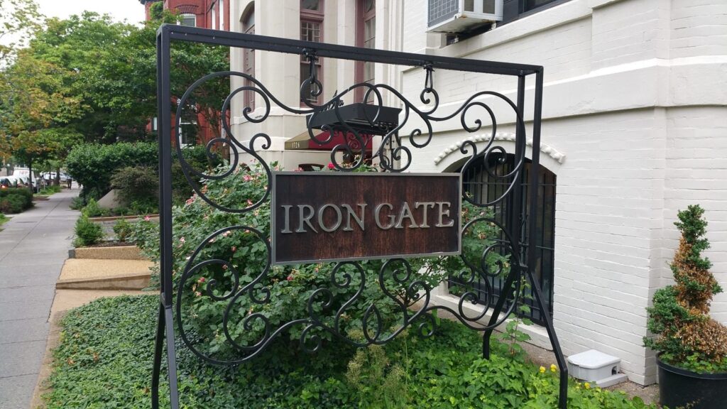 A decorative wrought iron sign with the words Iron Gate stands in front of a white building surrounded by greenery and a sidewalk on the left.