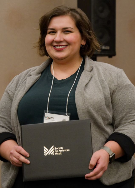 A person with short brown hair smiling and holding a black folder with the Society for American Music logo. They are wearing a gray blazer, dark top, and a name badge on a lanyard.