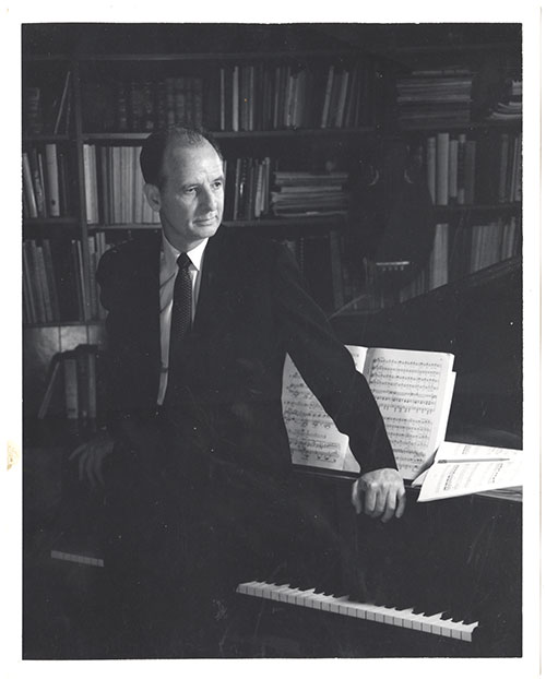 A man in a suit sits at a piano with sheet music, gazing to the left. A bookshelf filled with books is in the background. The image is in black and white.