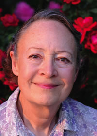 An older woman with short gray hair smiles gently, wearing a light purple patterned shirt, standing in front of vibrant pink and red flowers.