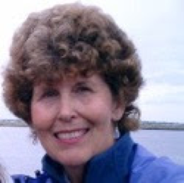 A woman with curly brown hair smiles while wearing a blue jacket, standing outdoors with water and sky in the background.