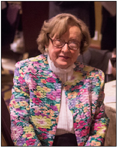 An elderly woman with glasses and short hair wears a colorful floral jacket and sits at a table indoors.