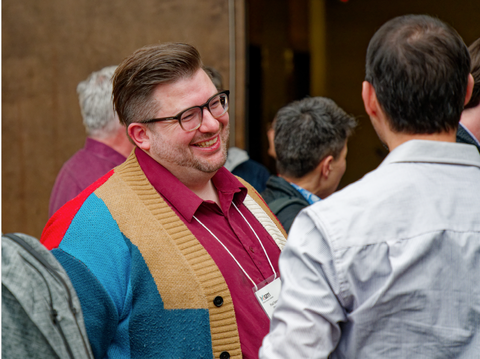 A smiling man in glasses and a colorful cardigan talks to another person at a social event, with others conversing in the background.