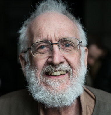 An elderly man with white hair, a full white beard, and glasses smiles warmly at the camera. He is wearing a brown shirt and is photographed against a dark, blurred background.