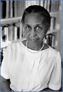 Black-and-white photo of an older woman wearing a white blouse and pearl necklace, standing in front of a blurred bookshelf, looking at the camera with a slight smile.