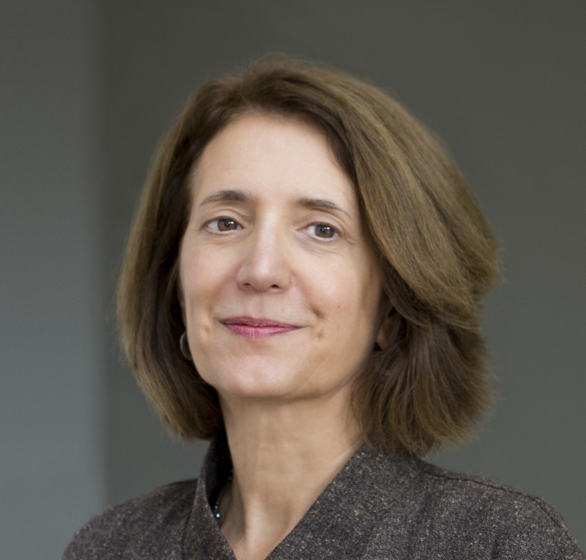 A woman with shoulder-length brown hair wearing a dark textured jacket and black top, standing against a plain gray background, looking at the camera with a slight smile.
