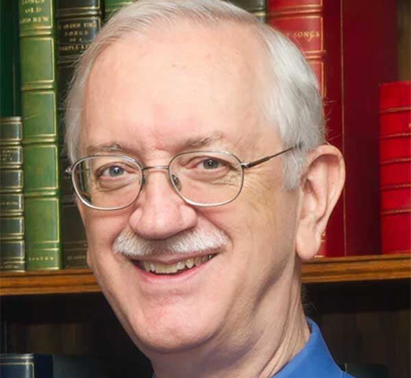 Smiling older man with glasses and white hair, wearing a blue shirt, standing in front of bookshelves filled with colorful books.